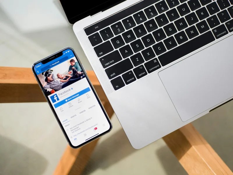 Facebook page on a mobile phone next to a MacBook.