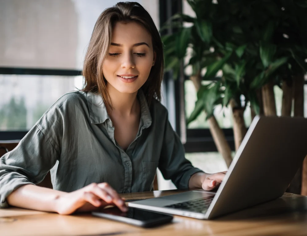Woman sat at a desk looking at her phone next to her laptop